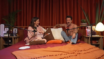 Roger González, sin camisa y en jeans, junto a una mujer con ropa rosa, ambos sentados en una cama con micrófonos y almohadas de "enKamados", frente a cortinas rojas