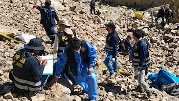 Agentes policiales y personal forense trabajan en un terreno rocoso. Una persona con traje azul, otra escribe en un portapapeles. Cinta policial amarilla en el suelo