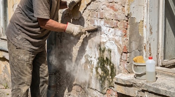 Persona con guantes cepilla una pared antigua y desmoronada para remover salitre y moho, levantando polvo. Cerca, un cubo con esponja y botella de limpiador.
