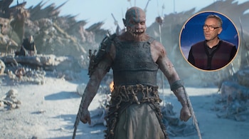 Un monstruo de piel rojiza, marcas azules y dientes afilados, vestido con armadura, en un paisaje desolado. En la esquina superior derecha, una imagen circular del director Simon McQuoid.