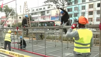 Varios obreros con cascos y chalecos arman estructuras metálicas de estrados en una avenida. Hay un autobús, edificios y cinta de seguridad