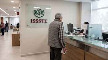 Un hombre de espaldas espera frente a una ventanilla con mampara de cristal en una oficina con el logotipo del ISSSTE.