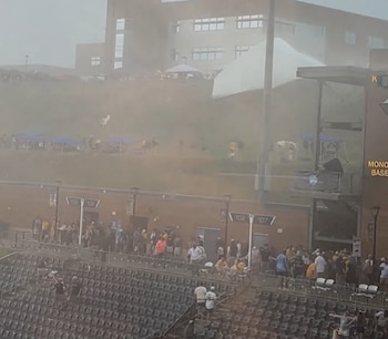 Vista de un estadio cubierto por una densa nube de polvo y escombros levantados por vientos fuertes, con personas dispersas en las gradas y una colina