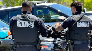 Dos agentes de policía de espaldas, con chalecos que dicen "POLICE", están junto a un vehículo policial oscuro y botellas de bebidas