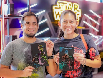 Hombre y mujer sonríen mientras sostienen dibujos de arte para rascar de Darth Vader y el Conde Dooku, con un letrero de neón de Star Wars de fondo