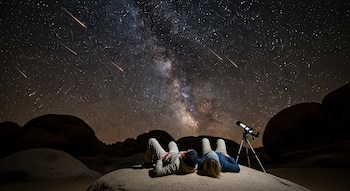 Dos personas tumbadas en una roca observando un cielo nocturno oscuro, lleno de estrellas, la Vía Láctea y varios meteoros; un telescopio está a su lado.