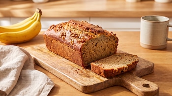 Pan de plátano con nueces en una tabla de madera, una rebanada cortada, un racimo de plátanos amarillos, un paño de tela y una taza.