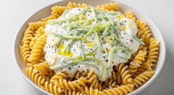 Plato blanco con fusilli dorados, salsa blanca cremosa, tiras de pepino verde, trozos de ajo, granos de pimienta negra y gotas de aceite de oliva.