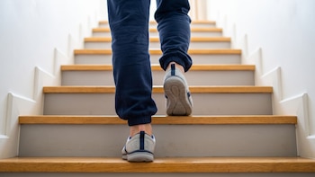 Pies y piernas de una persona con pantalón azul y zapatillas grises que sube por una escalera de madera