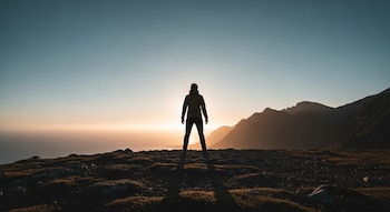 Silueta de una persona de pie en una cima rocosa, frente a un vasto horizonte con montañas y el mar bajo un cielo que transiciona del azul al dorado al amanecer.