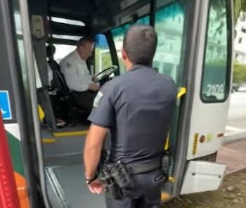 Un oficial de policía con el uniforme oscuro de espaldas, entrando a un autobús donde se ve al conductor sentado al volante