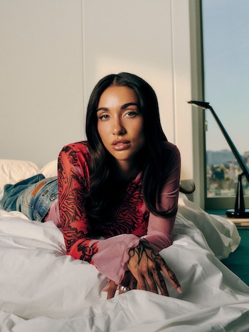 María Becerra yace boca abajo en una cama con sábanas blancas, mirando a la cámara. Viste una blusa estampada roja y rosa. Su mano izquierda está tatuada