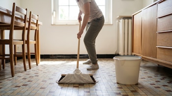 Se observa a una persona trapeando un piso de mosaico con una escoba cubierta por una jerga limpia, junto a una cubeta de plástico en un hogar ordenado.