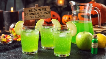 Tres vasos con bebida verde, hielo y rodajas de manzana, una jarra, una manzana verde, un cartel de madera y calabazas de Halloween.