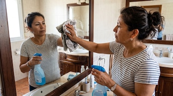 Una mujer sostiene un frasco limpiador azul mientras pasa un papel de periódico arrugado sobre el espejo de un baño.