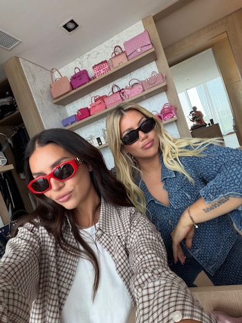 Dos mujeres con gafas de sol posan frente a una estantería de madera con varios bolsos de tonos rosados