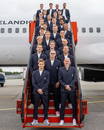 El plantel de fútbol de Noruega posa en fila sobre una escalera naranja y negra frente a un avión blanco. Todos visten trajes azules y camisetas blancas