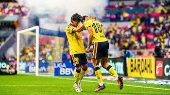 Dos jugadores de fútbol del Club América con uniformes amarillos se abrazan en el campo. Se ven espectadores, un arco de portería y vallas publicitarias.