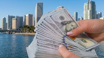 Una mano sujeta un fajo de billetes de cien dólares, con el horizonte de rascacielos de Miami, palmeras y el agua del río en el fondo bajo un cielo despejado.
