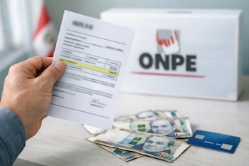 Mano sostiene multa impresa. Billetes de 100 soles peruanos y tarjeta de débito sobre mesa, con urna ONPE y bandera peruana desenfocadas al fondo.