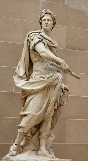 Estatua de mármol de cuerpo entero de un hombre con corona de laurel, túnica, coraza, capa, sandalias, un bastón y un escudo, sobre fondo de pared de piedra