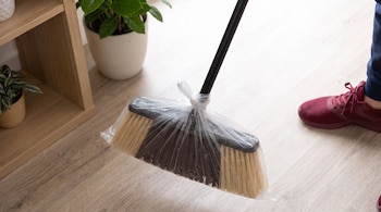 Una escoba con cerdas marrones y amarillas cubierta con una bolsa de nylon transparente sobre un suelo de madera, junto a una pierna y zapatilla guinda.