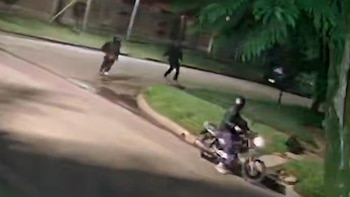 Grabación granulada de cámara de seguridad nocturna mostrando a una persona en una motocicleta y dos figuras oscuras corriendo por una calle y acera con árboles