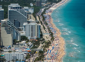 Vista aérea de una larga playa de arena dorada llena de personas y sombrillas de colores, junto a una carretera con tráfico y edificios altos a lo largo de la costa