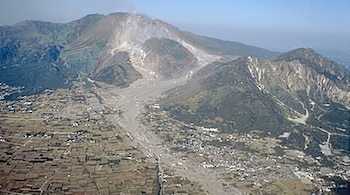 Vista aérea de una montaña volcánica con humo en la cima y un cauce de ceniza gris que atraviesa campos de cultivo y casas.