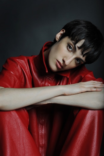 Retrato de mujer joven con pelo corto oscuro, vestida con atuendo de cuero rojo, brazos cruzados, sobre fondo gris oscuro