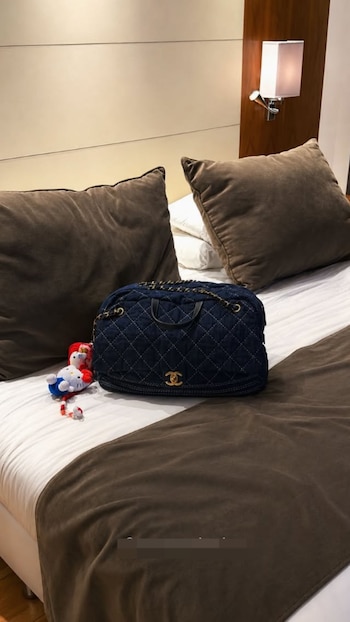 Una cartera Chanel azul marino acolchada y dos pequeños peluches de Hello Kitty están sobre una cama con sábanas blancas y almohadas marrones en un hotel