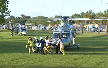 Personal de emergencia traslada un paciente en camilla a un helicóptero médico gris y verde en campo de hierba. Otro helicóptero está detrás