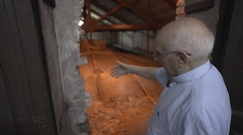 Un sacerdote con anteojos y camisa clerical señala con la mano abierta hacia un espacio iluminado detrás de una pared de piedra.