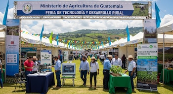 Una feria al aire libre con carpas blancas y personas explorando exposiciones de tecnología agrícola bajo un gran cartel del Ministerio de Agricultura de Guatemala.