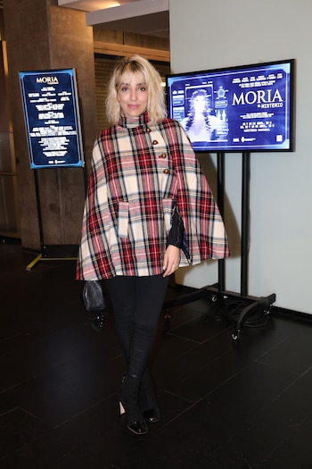 Mujer rubia de cabello corto posa de pie, con poncho a cuadros rojo, blanco y negro, pantalones y botas oscuras. Detrás, dos pantallas azules con texto