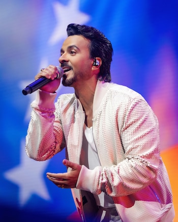 Luis Fonsi sostiene un micrófono mientras canta en un escenario iluminado con luces azules y rojas