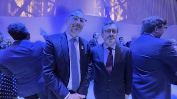 Dos hombres con traje y corbata posan frente a la cámara en un salón iluminado con luz azul junto a otros asistentes
