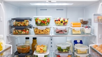 Interior de un refrigerador iluminado con recipientes transparentes que contienen guisados, ensaladas, frutas y quesos en sus estantes.