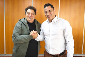 Dos hombres sonrientes, uno con sudadera oscura y perilla, el otro con camisa blanca, estrechan sus manos frente a una pared de paneles de madera