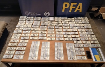 Billetes de cien dólares apilados ordenadamente sobre una mesa de madera, con dos pancartas azules de la Policía Federal Argentina (PFA) al fondo