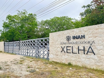 Un cartel de piedra con el texto "INAH ZONA ARQUEOLÓGICA XELHÁ" y una reja metálica. Al fondo se ven árboles y el cielo nublado