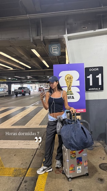 Mujer con gorra y teléfono junto a carrito con equipaje. Detrás, un cartel del trofeo del Mundial FIFA y señal de arribos en un estacionamiento