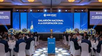 Salón de eventos con mesas redondas y personas sentadas, un escenario con pantallas azules, un trofeo, arreglos florales y la bandera de Guatemala.