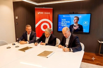 Tres hombres en traje sentados en una mesa blanca, dos de ellos firmando documentos. Detrás, un cartel de BID Invest y una pantalla con publicidad de Banco Macro