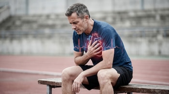 Hombre atlético de mediana edad, de perfil, sentado en un banco con ropa deportiva, tocándose el pecho. Un gráfico rojo y azul de un corazón y un ECG se superpone en su torso.