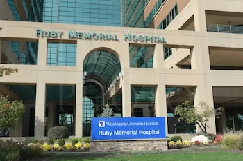 Fachada del edificio Ruby Memorial Hospital con ventanales de vidrio, un arco central y un letrero azul de entrada sobre el jardín