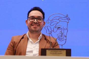 Un hombre con gafas y saco gris sostiene un trofeo metálico con una placa grabada frente a la cámara