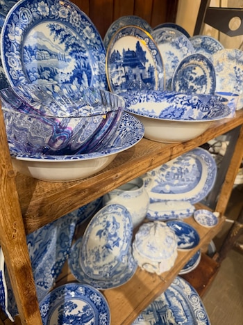 Estanterías de madera con una variada colección de platos, cuencos y piezas de cerámica decoradas en patrones azules y blancos, con algunos detalles dorados y cristalería