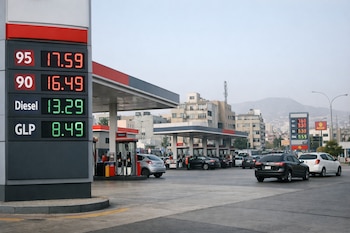 Primer plano de un poste con precios digitales de gasolina, diésel y GLP, y al fondo, una estación de servicio con surtidores, coches y edificios urbanos.