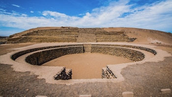 Gran estructura circular de piedra con un suelo de tierra clara en primer plano, y una pirámide escalonada de piedra y tierra al fondo bajo un cielo azul con nubes